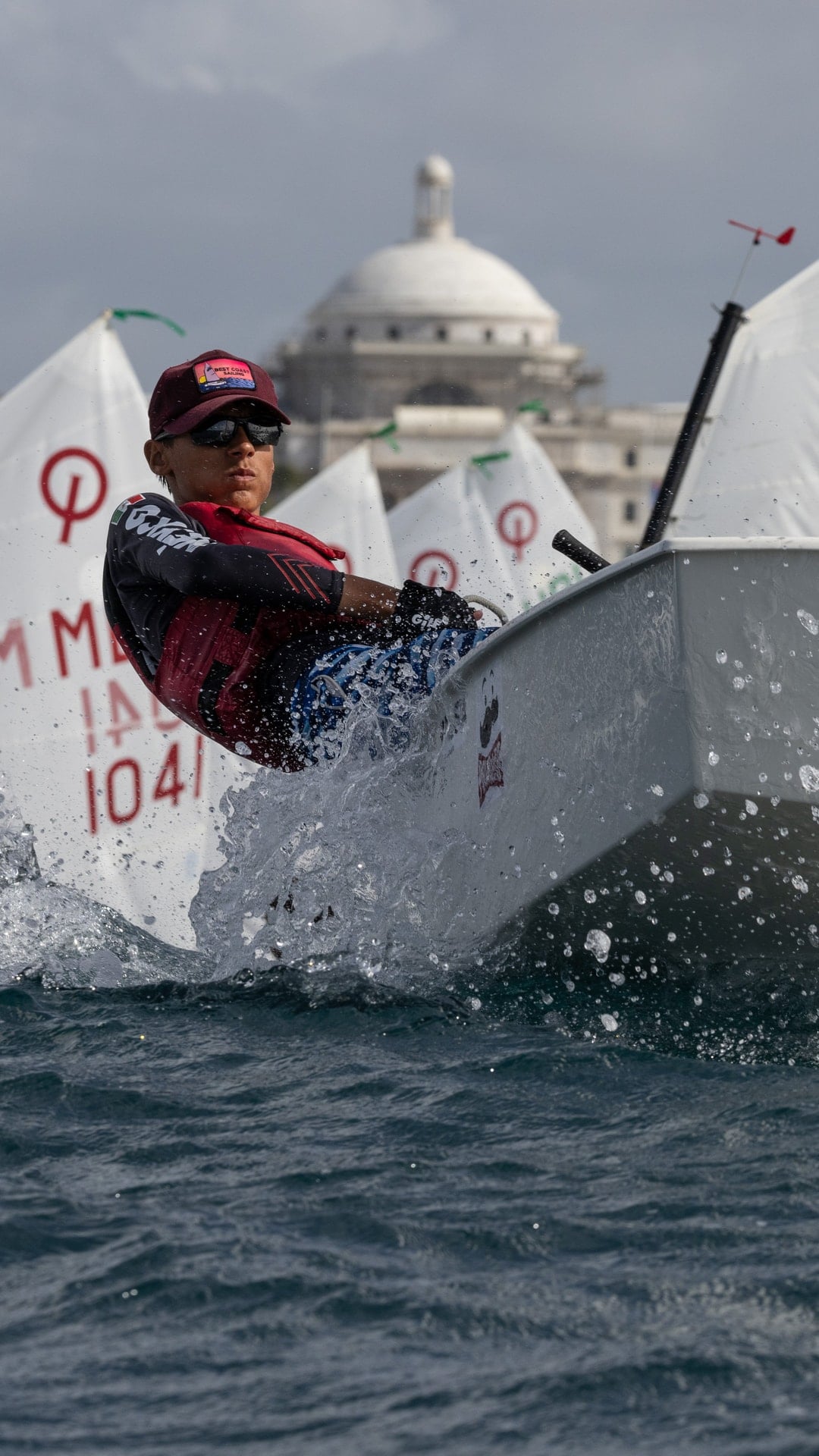 A young sailor hiking out as an Optimist dinghy punches through spray, with a fleet of sails behind